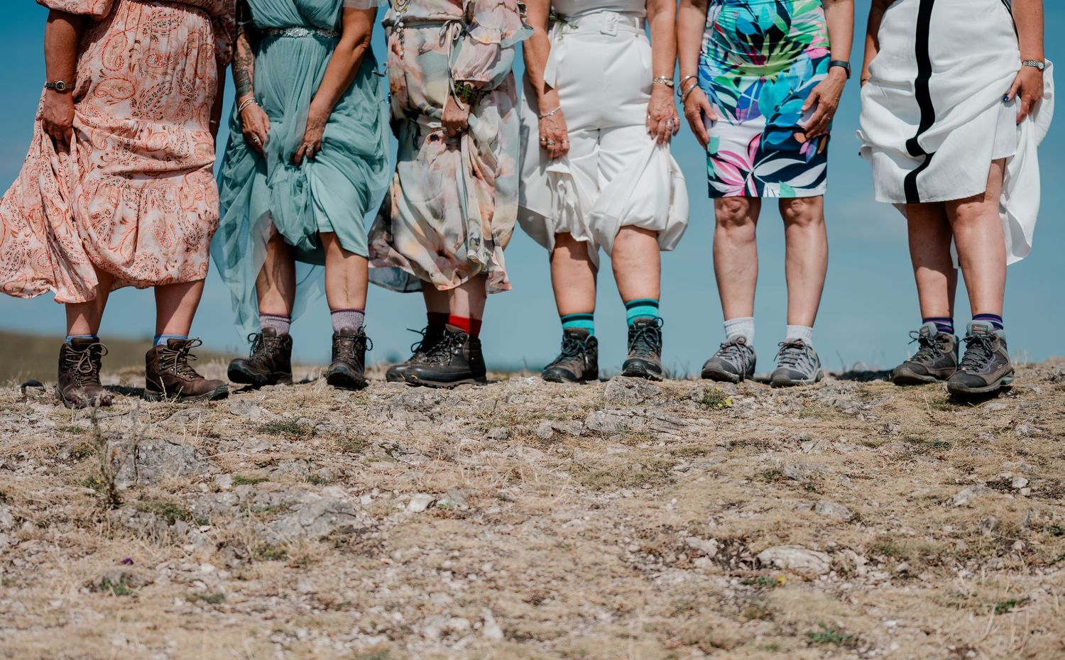 Group of wedding guests showing muddy boots and colorful socks standing on rocky terrain under blue sky.