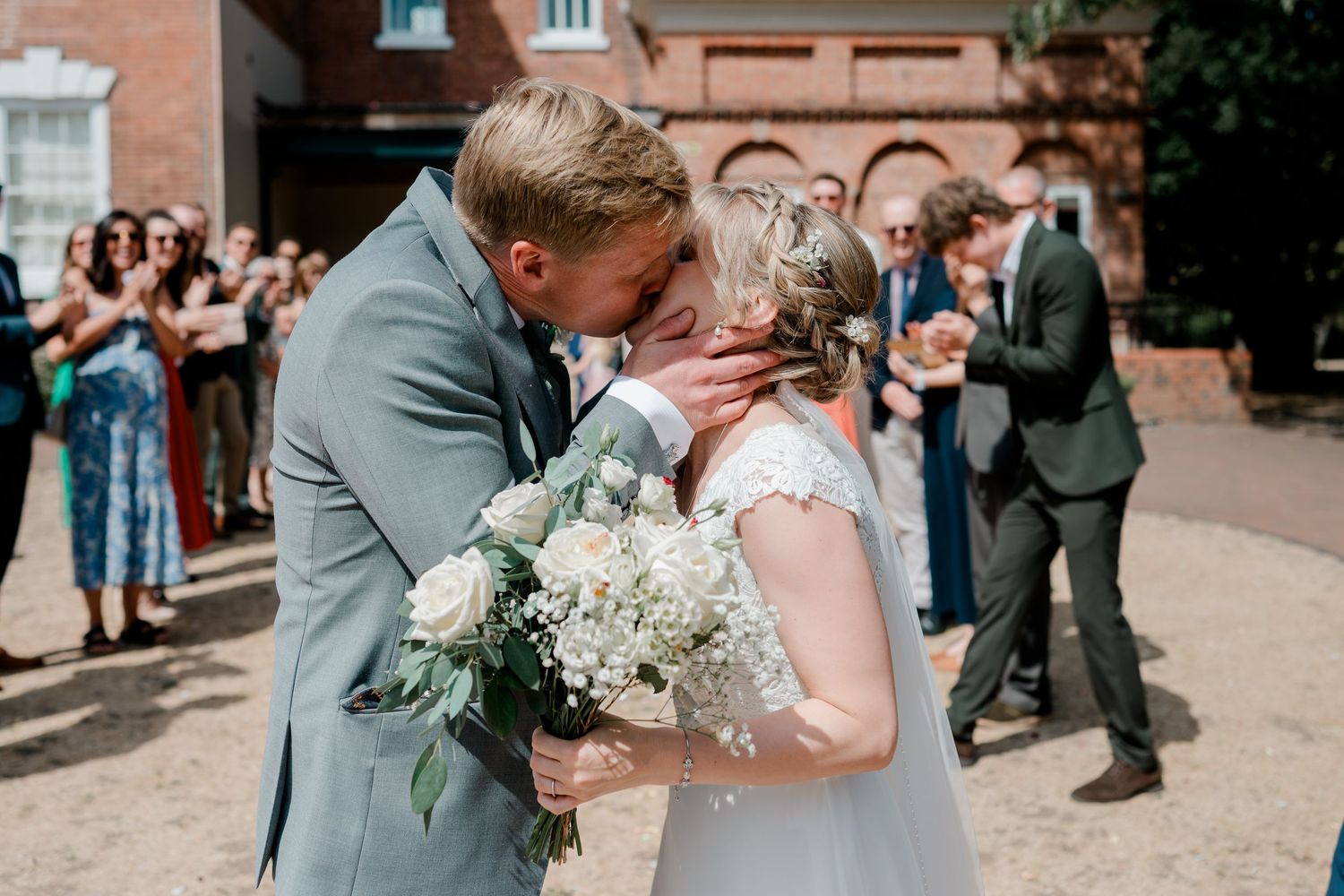 Newlywed couple sharing a first kiss outdoors after wedding ceremony, bride holding white floral bouquet with guests applauding.