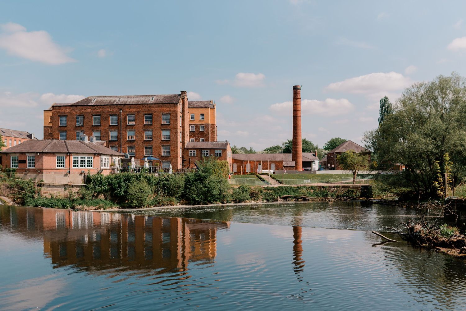 Stunning converted mill wedding venue reflected in a tranquil river with iconic chimney stack backdrop.