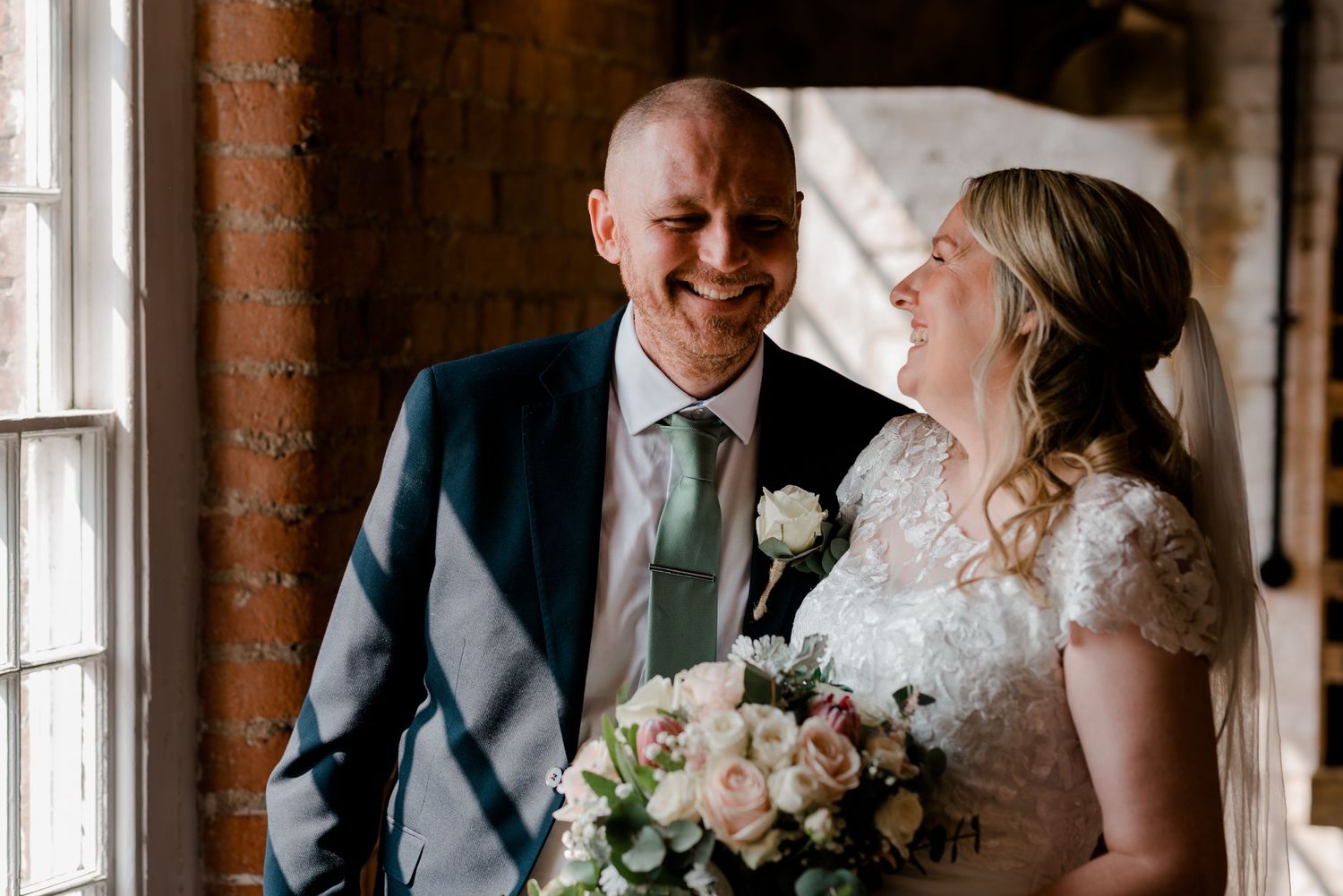 Laughing bride and bald groom share a joyful moment indoors beside a window with rustic brick wall.