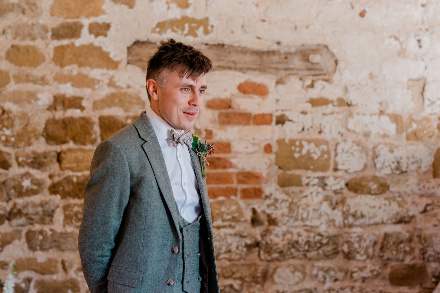 Wedding party members in grey suits pose against rustic exposed brick wall.