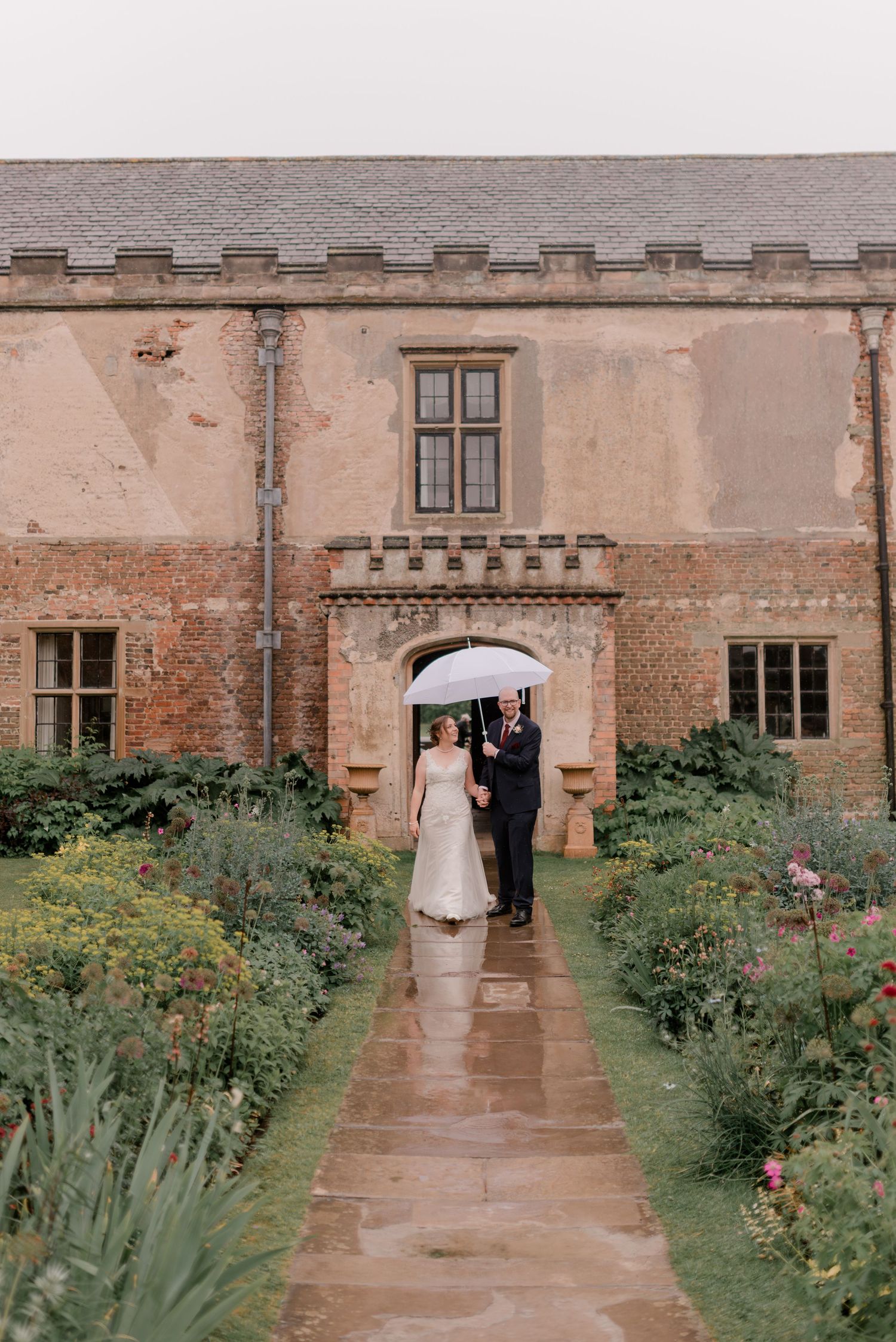 A couple stands in the stone archway of a historic manor house with beautiful garden paths and flower beds on a rainy day.