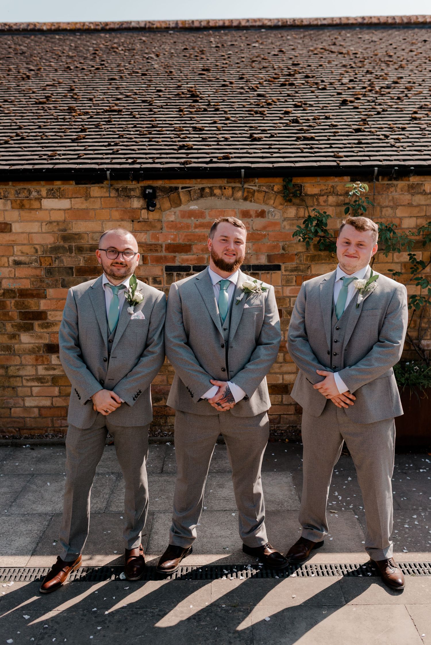 Groomsmen lined up against brick wall wearing matching gray suits in formal pose.