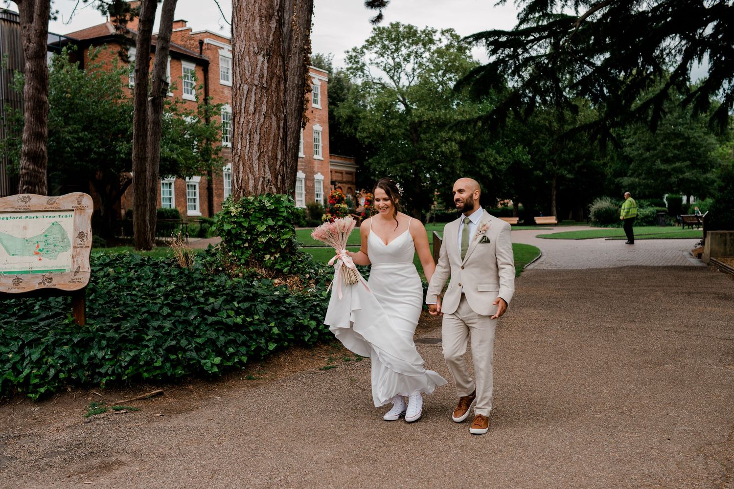 Bride and groom walking hand in hand outdoors at a garden venue, bride in white gown with bouquet, groom in beige suit.