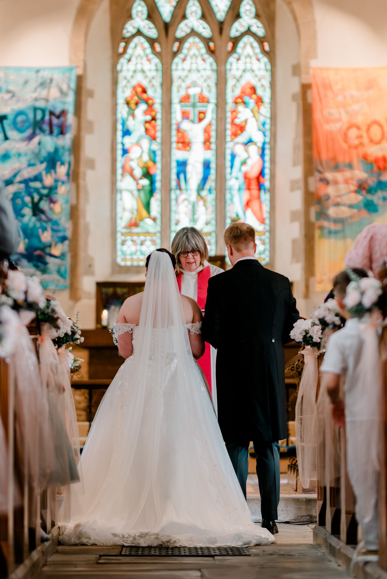 Bride and groom stand at church altar before officiant with stained glass windows during wedding ceremony.