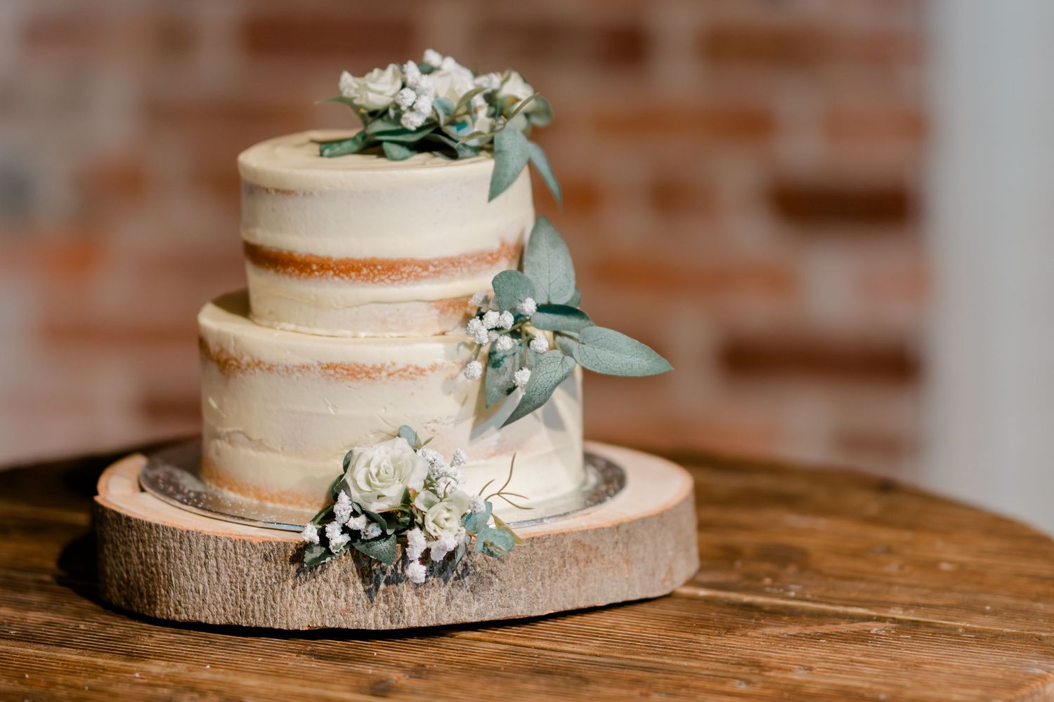 Two-tier naked wedding cake with white roses and eucalyptus on a rustic wood slice stand.