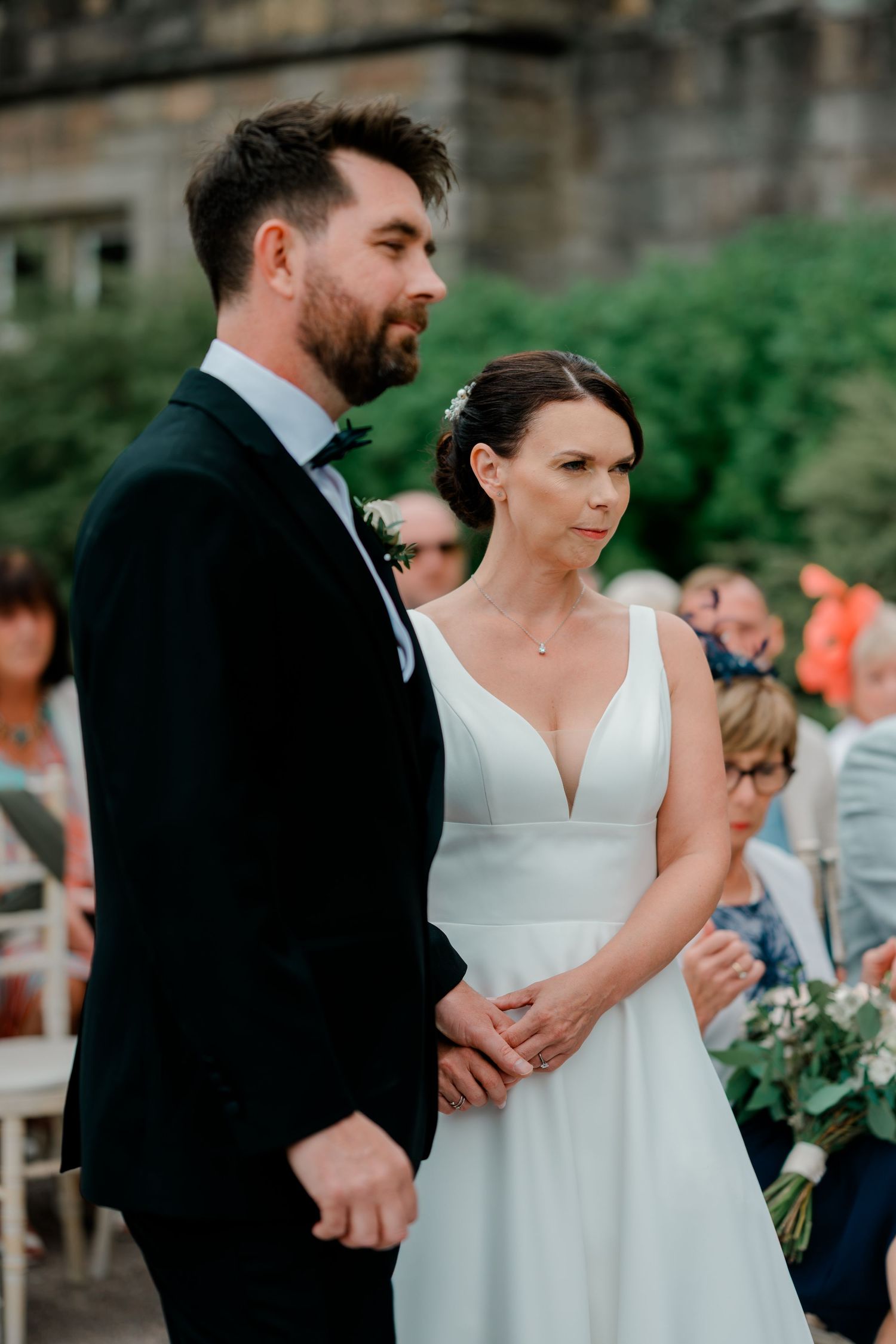 Bride in a white V-neck wedding dress holds hands with her groom in a dark suit during an outdoor ceremony as guests look on.