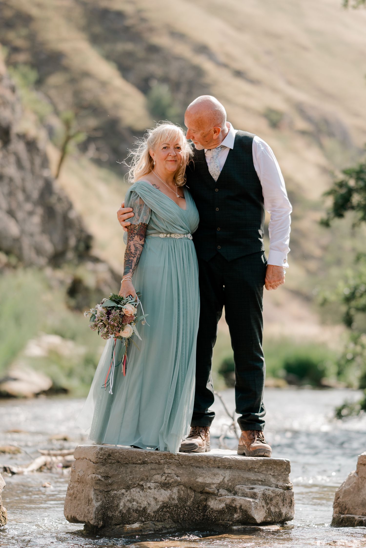 Elegant older couple in wedding attire standing on rocks by a mountain stream, sharing a tender romantic moment.