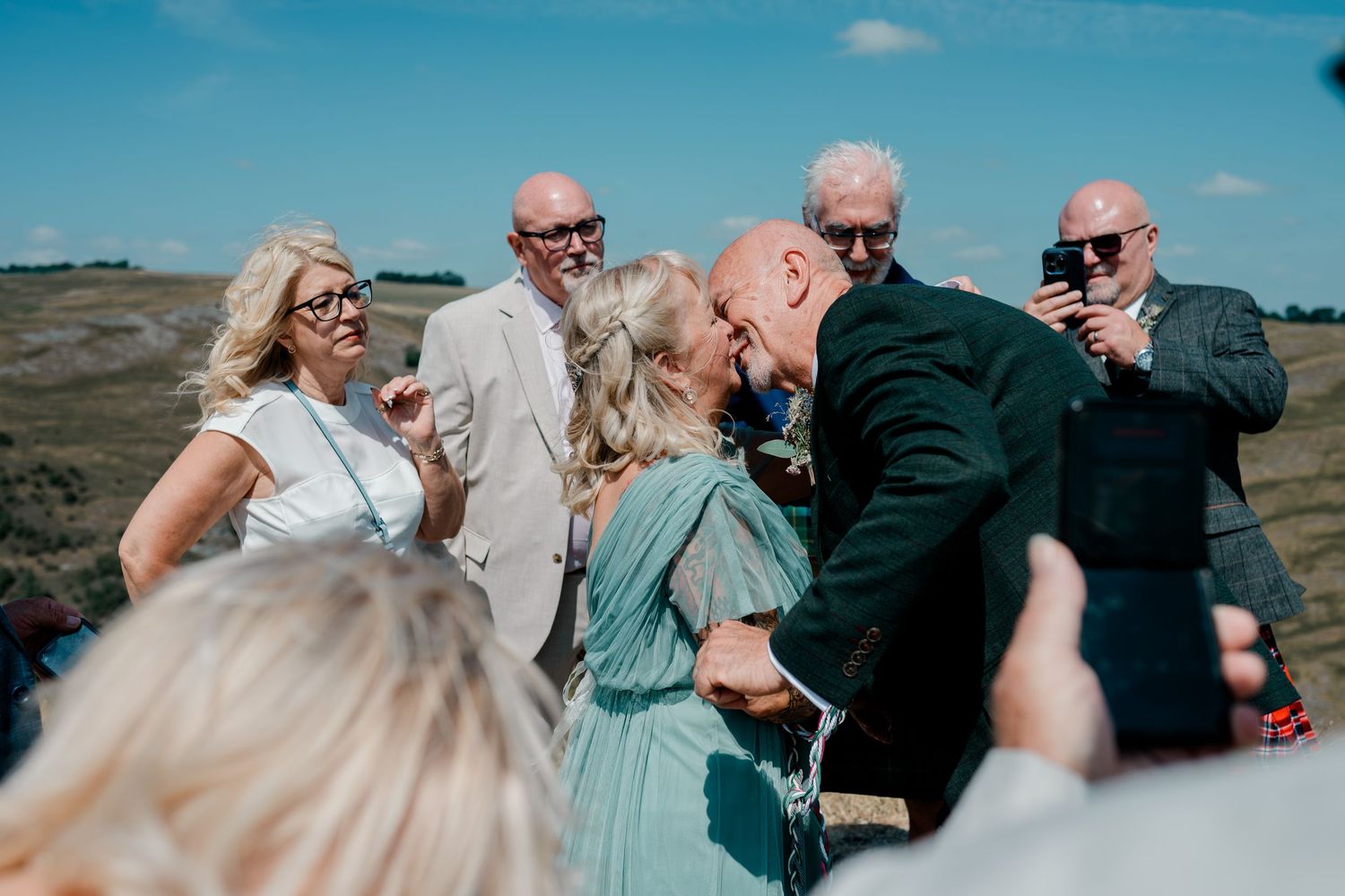 Bride and groom share a kiss surrounded by smiling wedding guests outdoors.