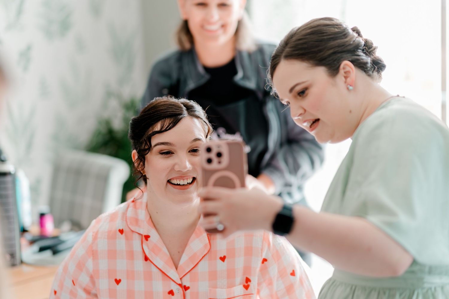 Bride in pink gingham pajamas laughing while viewing photos on a phone with bridesmaids during wedding prep.