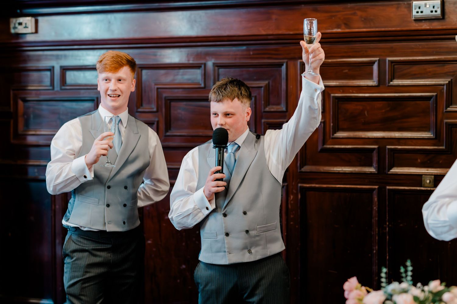 Two young groomsmen in grey vests delivering a wedding speech, one raising a glass in a toast.