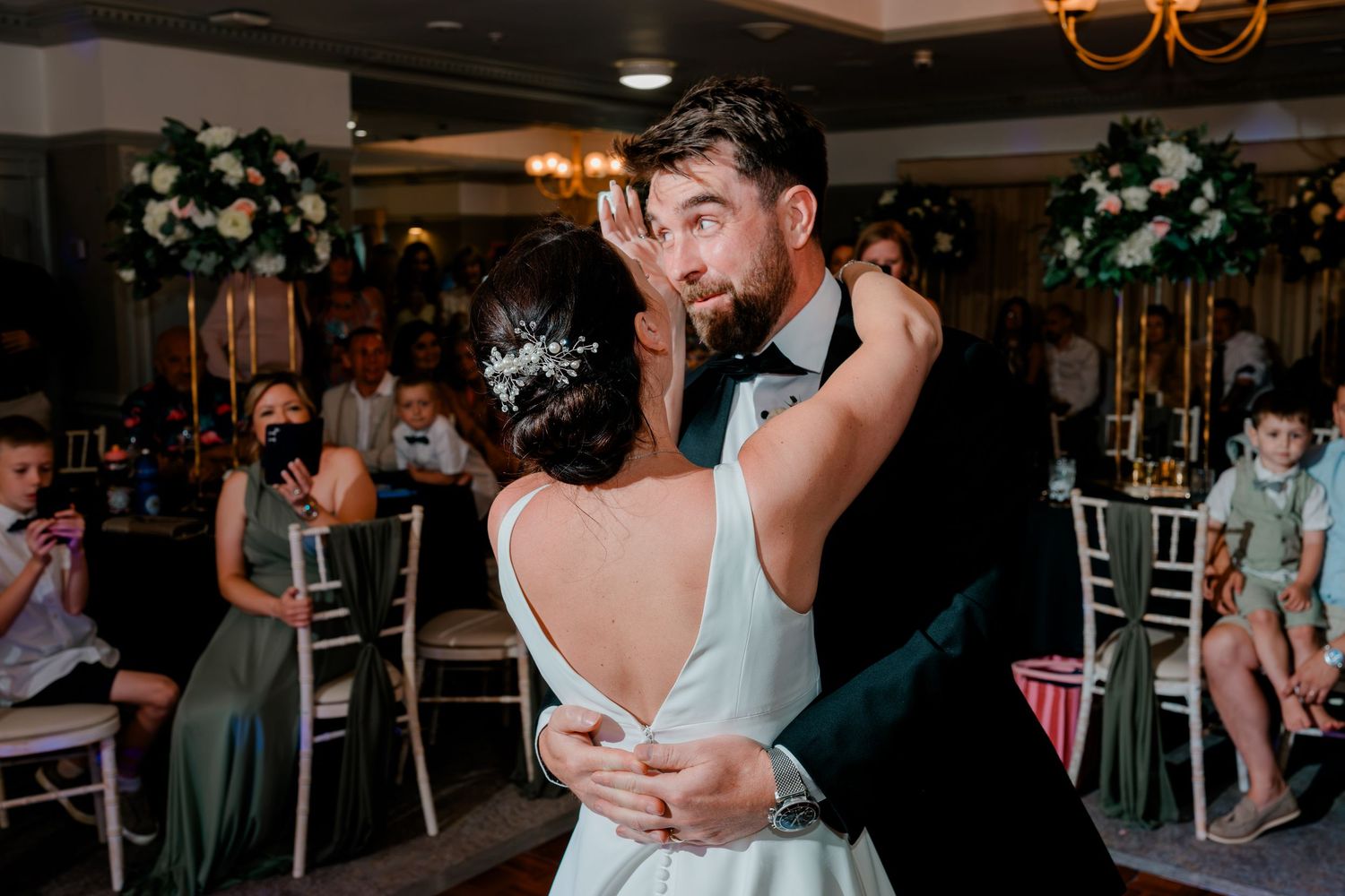 Bride in a white backless gown with a sparkling hair accessory shares a first dance with a smiling groom as wedding guests watch.