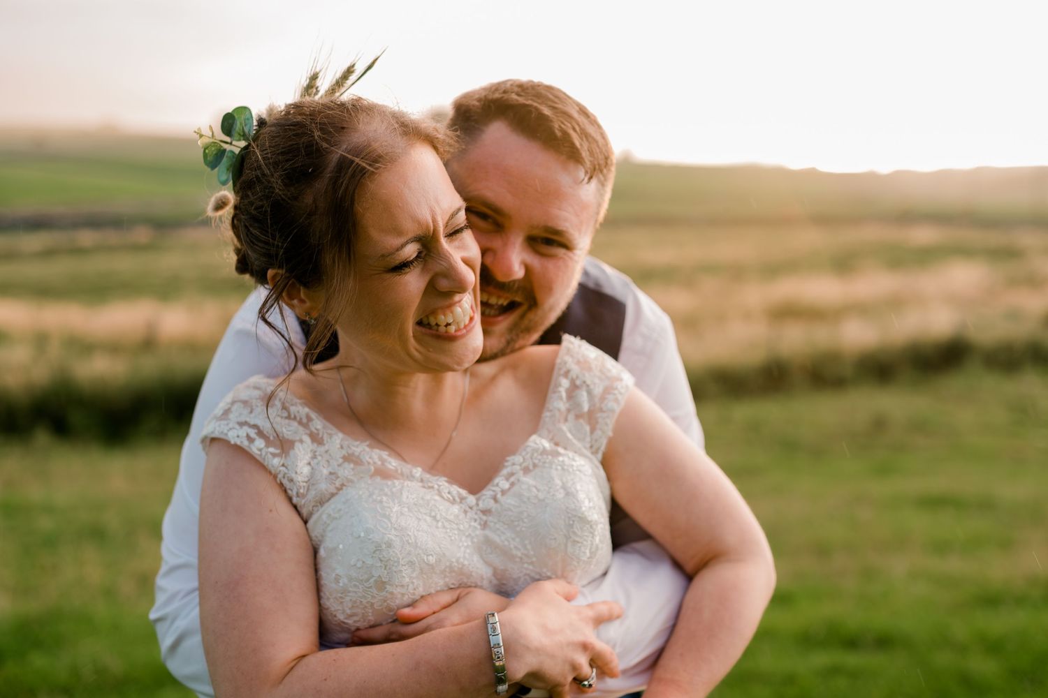 Groom hugging bride from behind as she laughs joyfully in a sunlit green field on their wedding day.