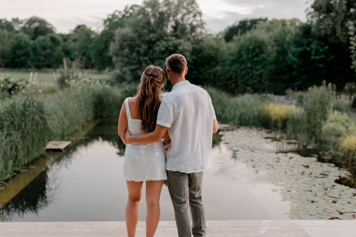 Couple in white standing arm in arm on a dock overlooking a tranquil lily pad pond.