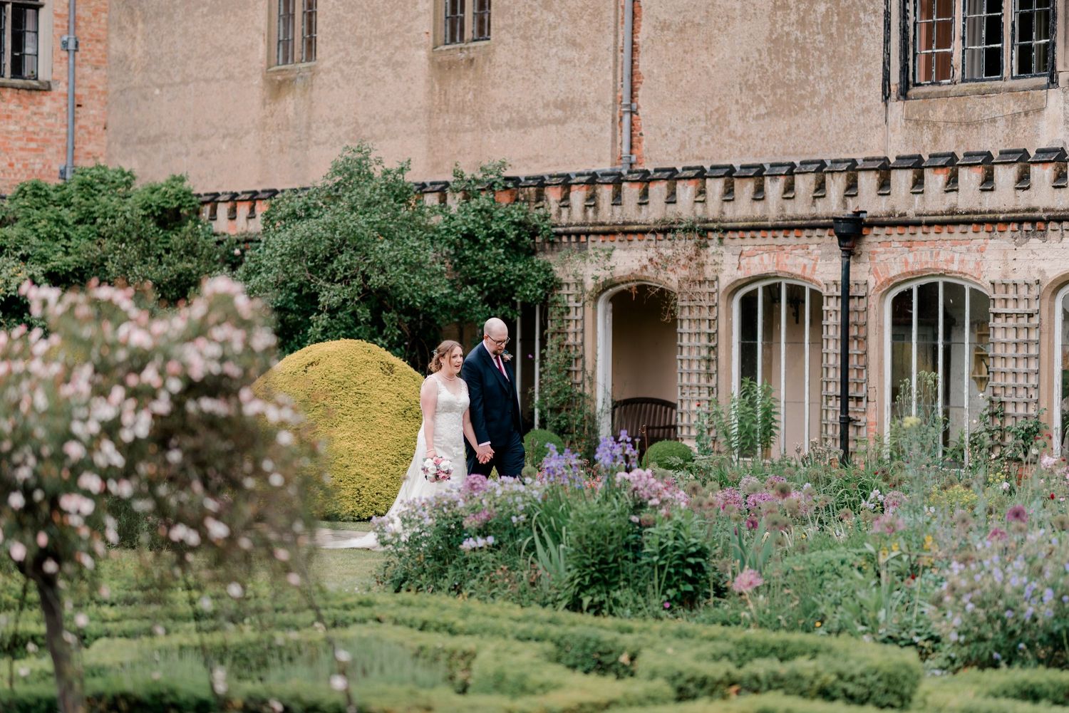 Elegant couple walks through stunning formal garden beside grand historic stone manor house.