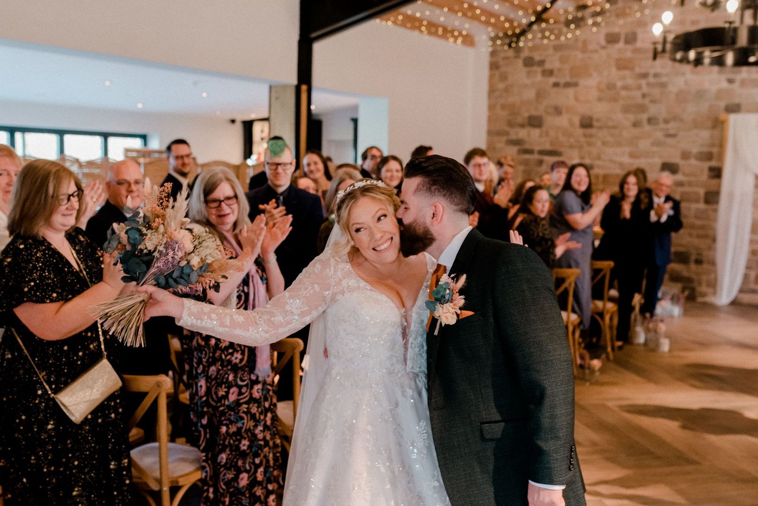 Joyful bride and groom walking back down the aisle greeted by cheering wedding guests in a stone barn venue.