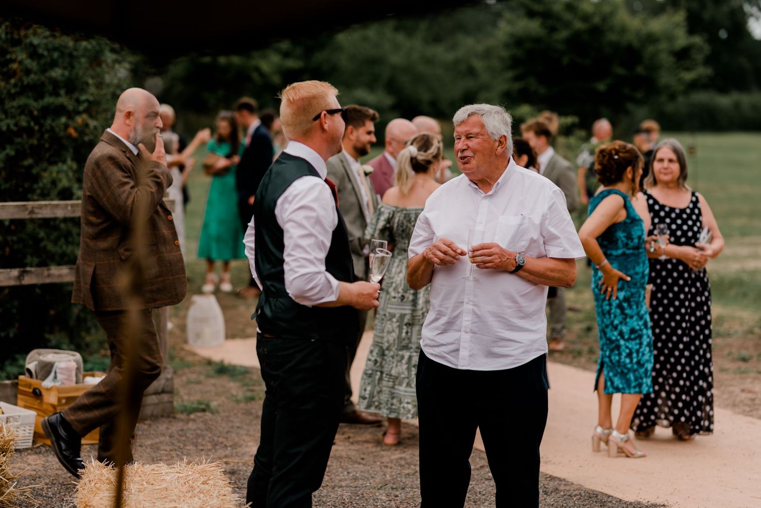 Guests enjoy casual conversation at an outdoor wedding reception with people in formal attire and summer dresses.