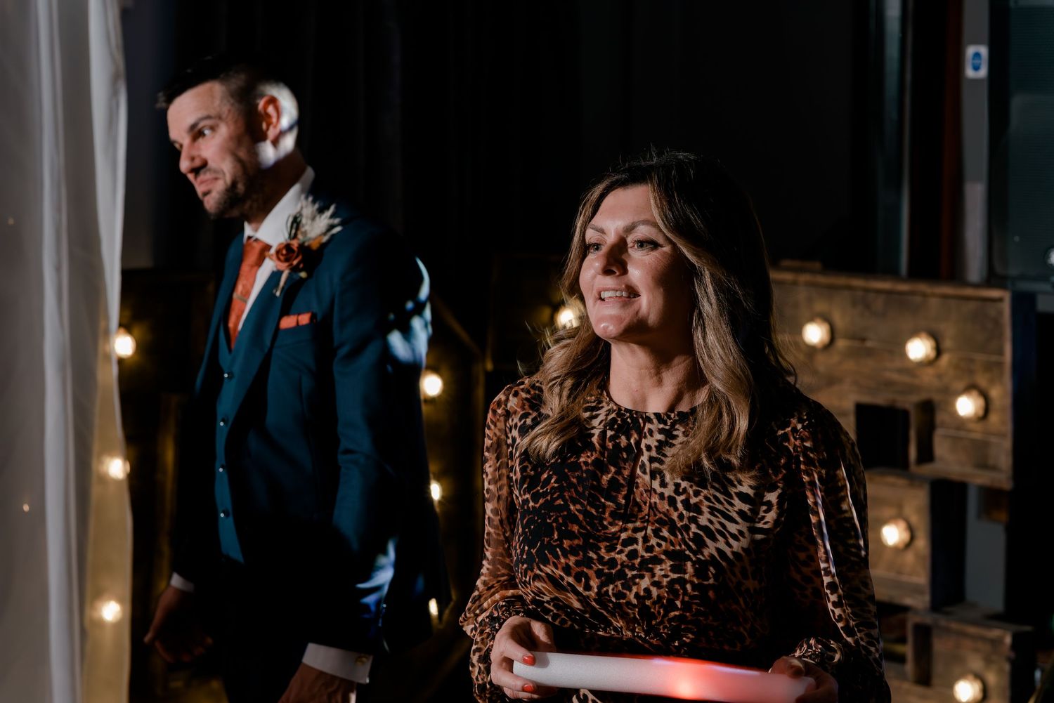 A couple stands in front of illuminated lights with the woman wearing a leopard print dress and the man in a suit.