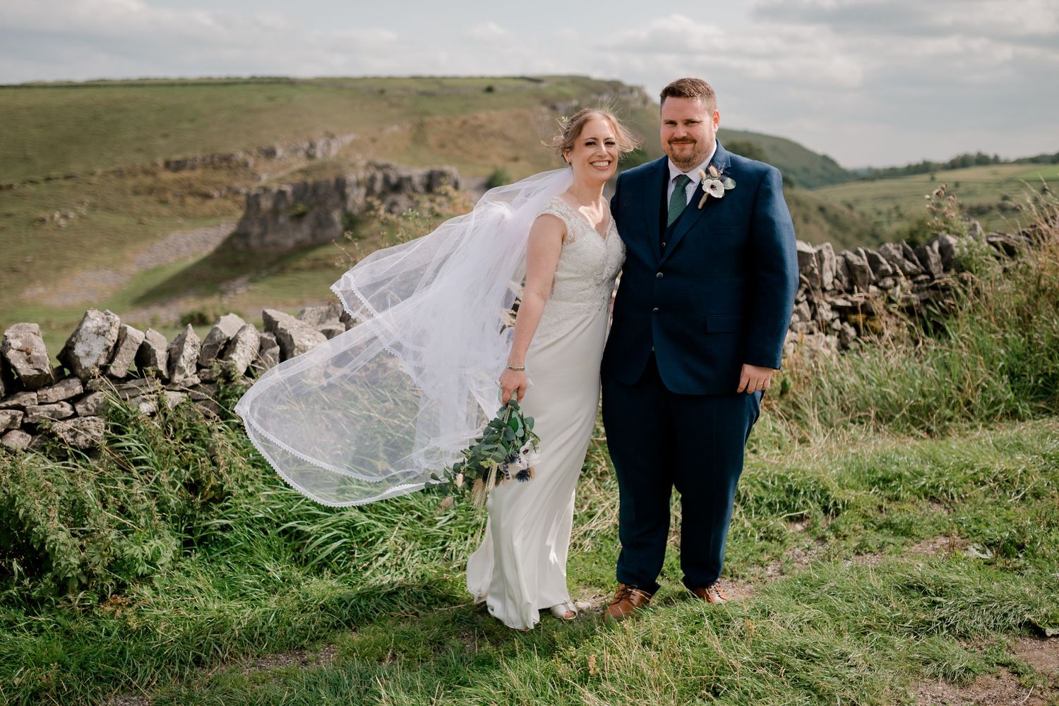 A couple poses for romantic wedding photos on a scenic hillside with stone walls and flowing white veil in the breeze.
