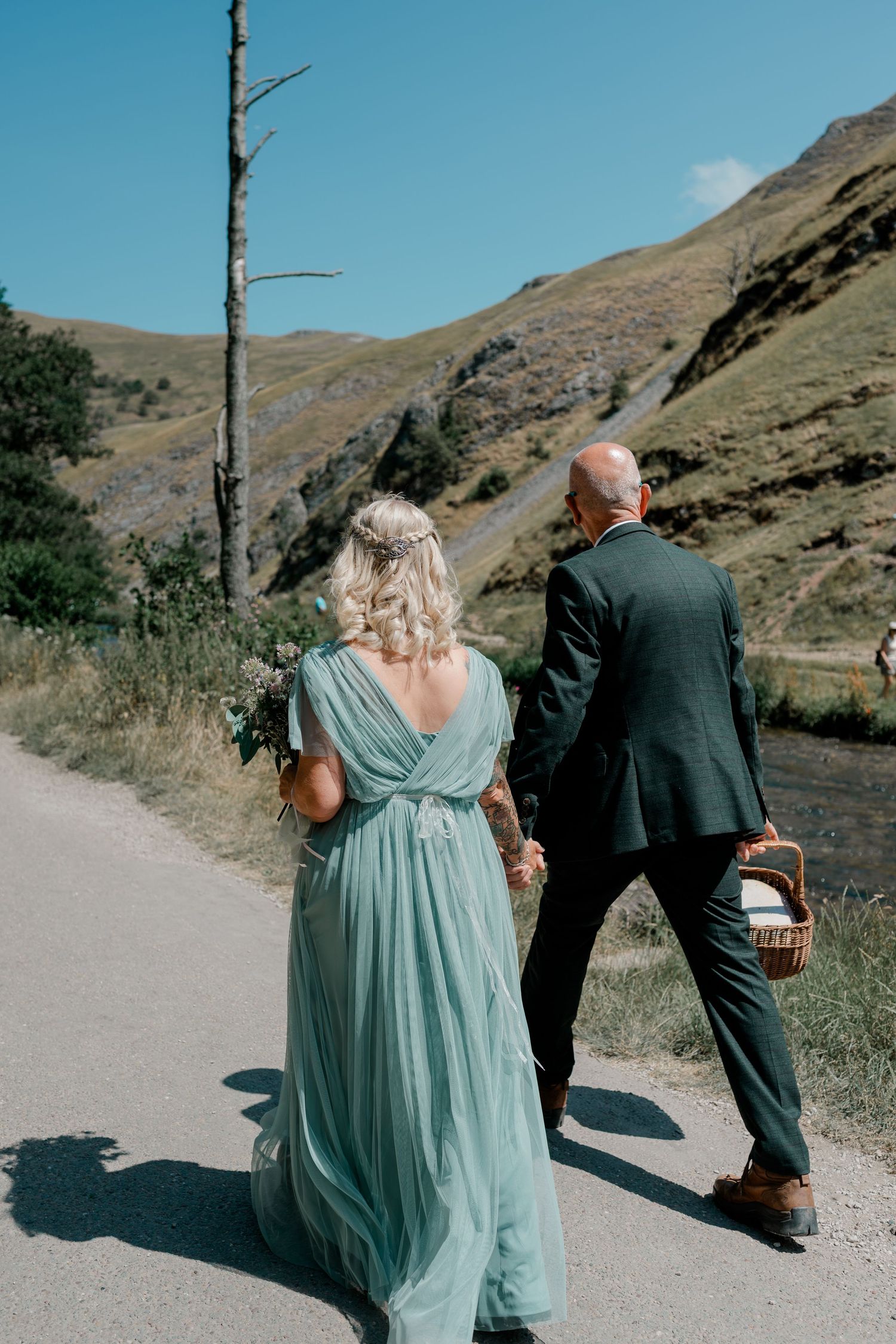 Bride in teal dress and groom in dark suit walk hand-in-hand along a scenic mountain road.