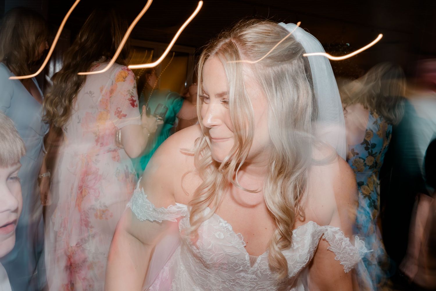 Bride in lace off-shoulder wedding dress and veil dancing joyfully at reception with motion blur effect.
