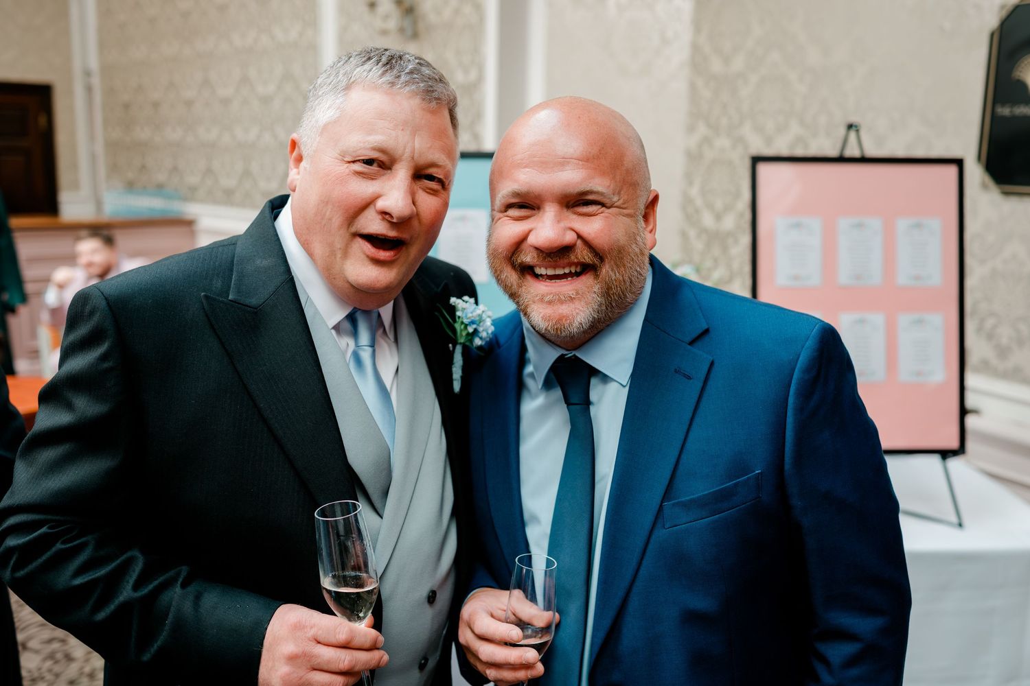 Two smartly dressed wedding guests smiling and holding champagne glasses at a reception.