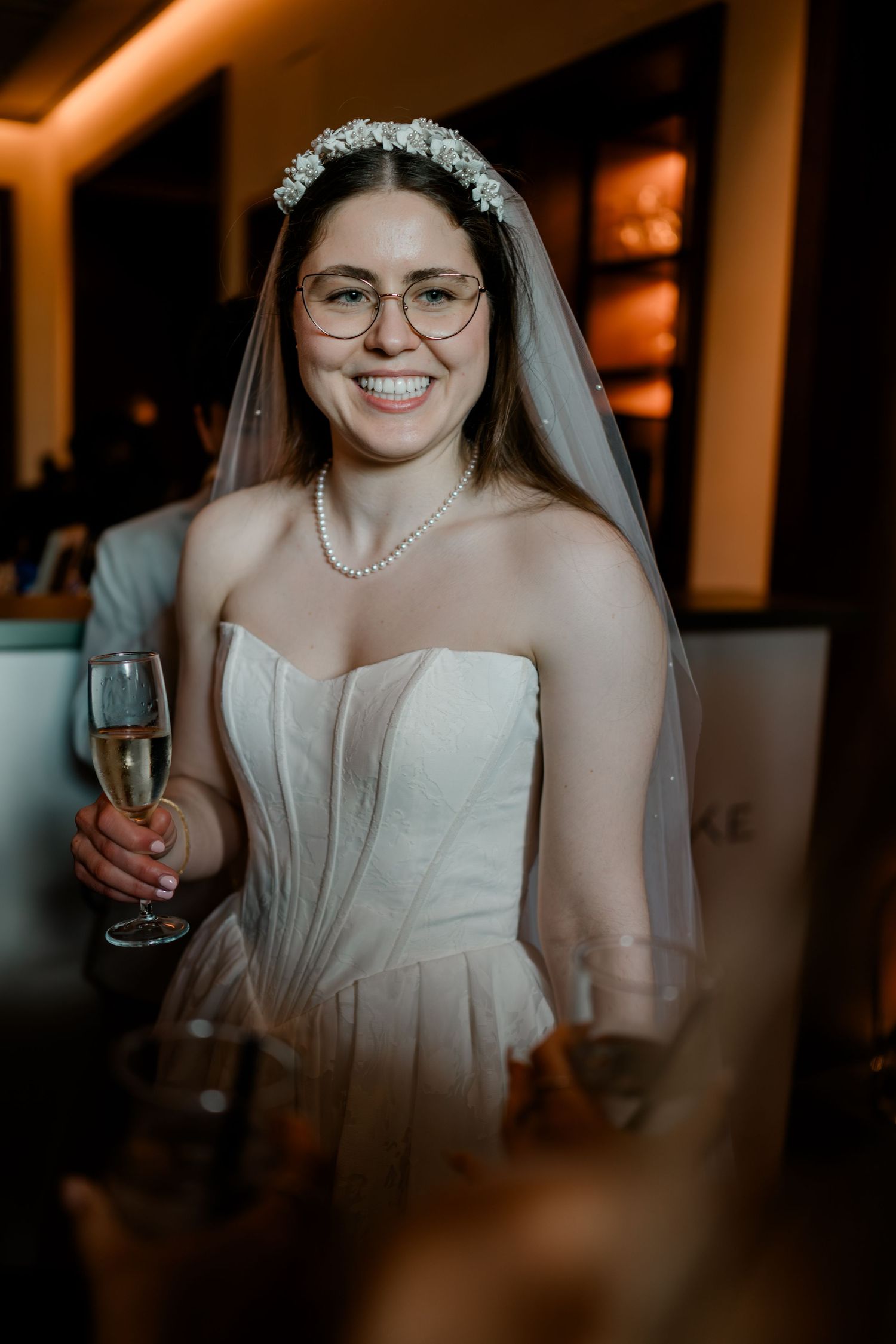 Smiling bride in strapless gown, pearl necklace, floral headband, and veil holding a champagne glass.