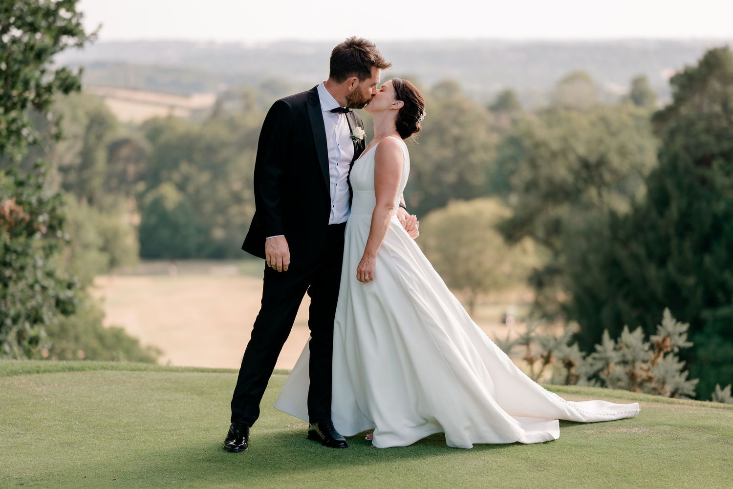 Bride and groom share a romantic kiss outdoors on a green lawn with scenic countryside views on their wedding day.