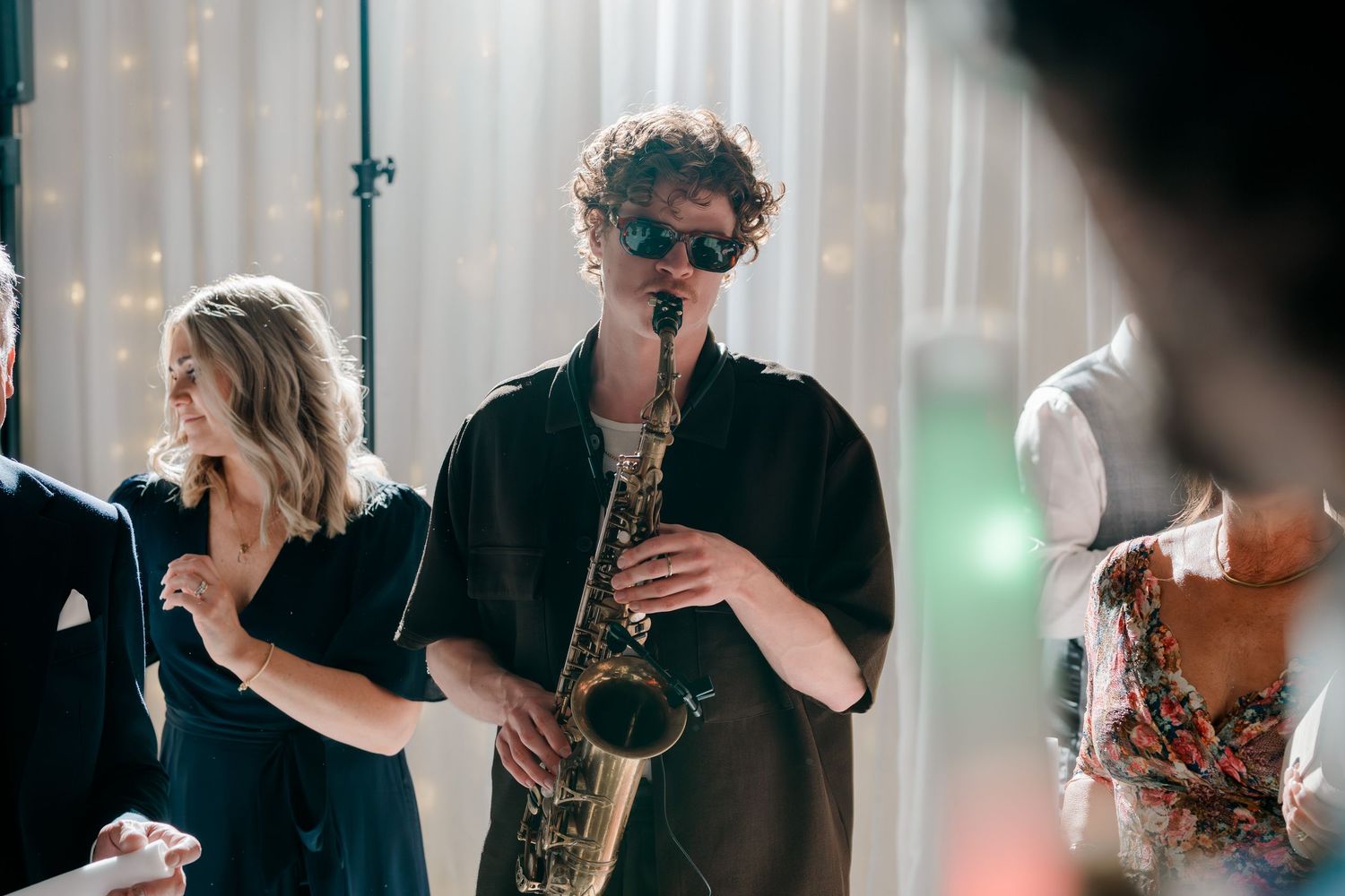 Saxophonist in black outfit performing live music at a wedding reception with fairy lights.