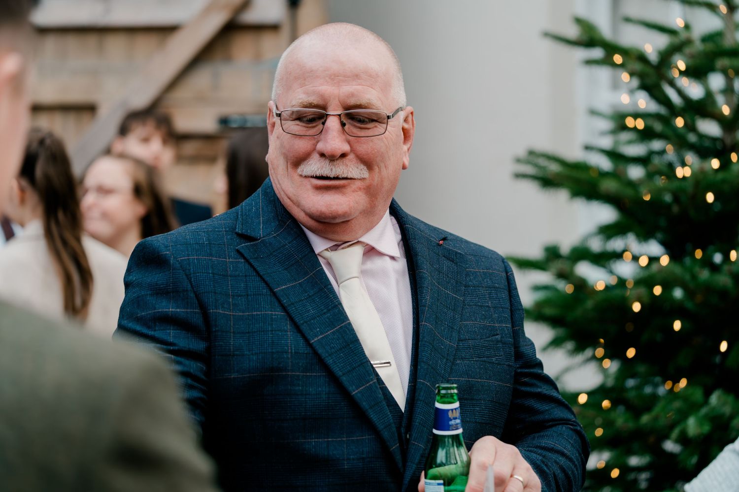 Distinguished male wedding guest in navy suit smiles holding green bottle at reception.