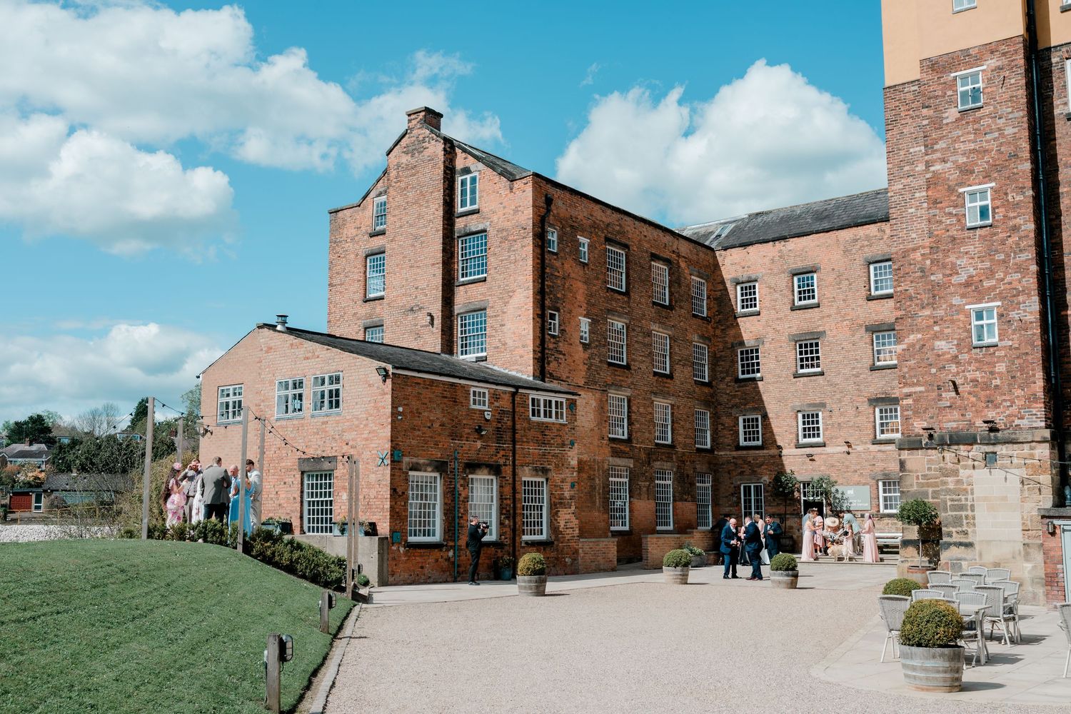 Historic red brick mill building with courtyard under blue sky, featuring Georgian architecture and visitors exploring the grounds.