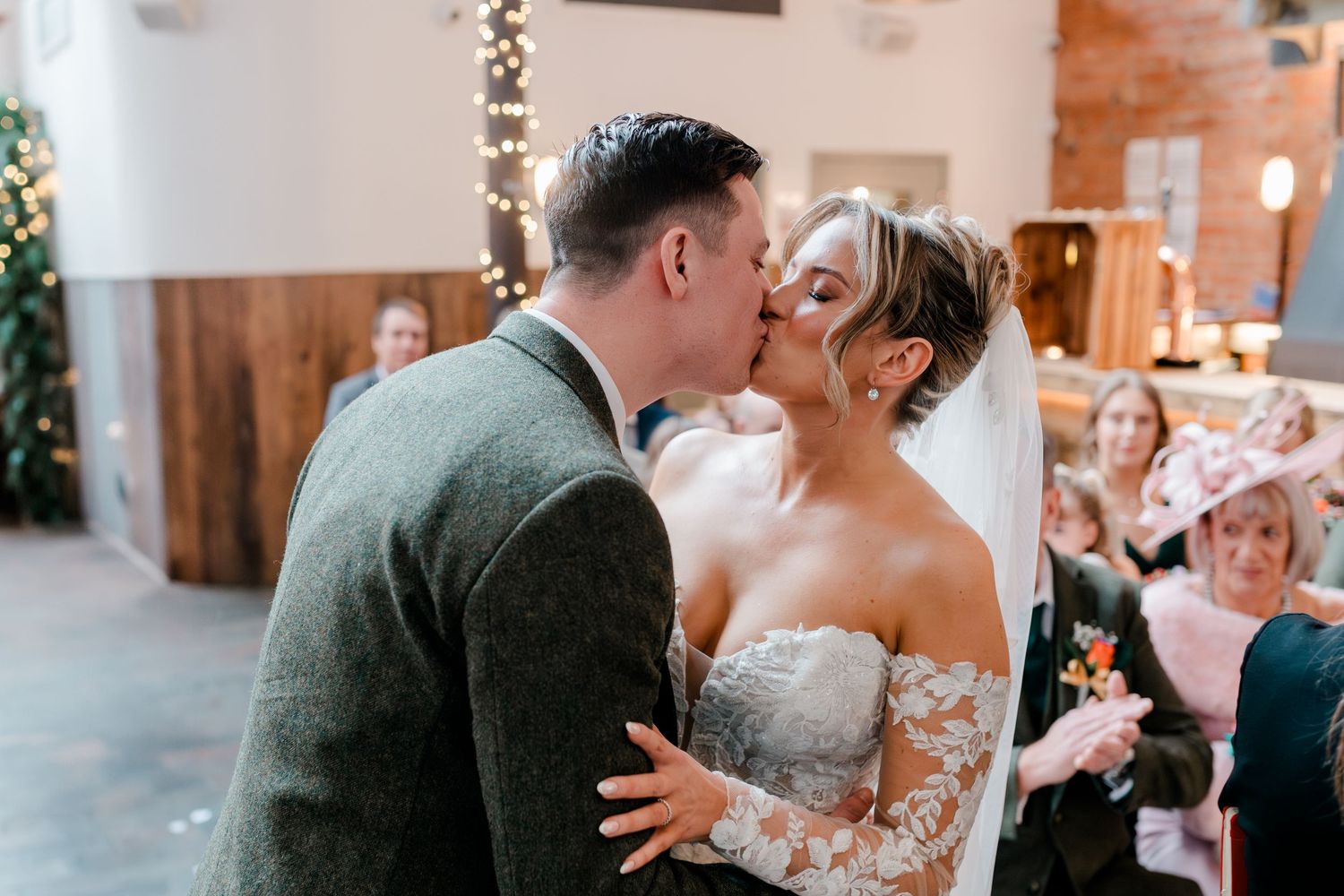 Bride and groom sharing first kiss at rustic indoor wedding ceremony with fairy lights.