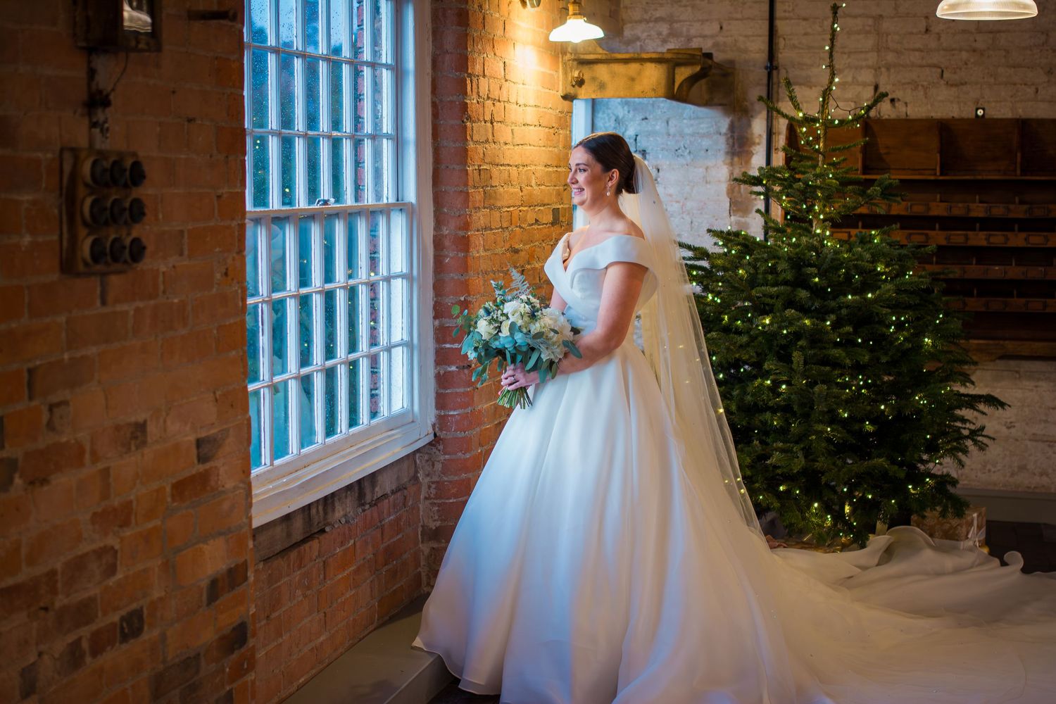 A white bridal gown displayed in a rustic brick venue with natural window light and a Christmas tree.