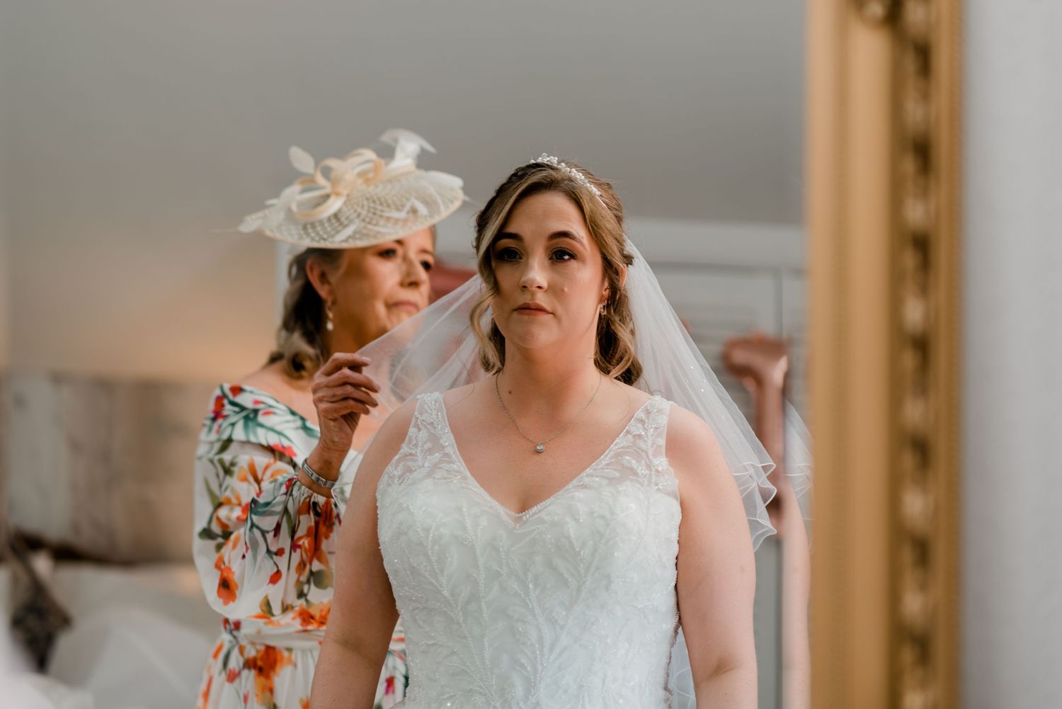 Two people getting ready for a wedding ceremony with someone adjusting a white veil and lace gown.