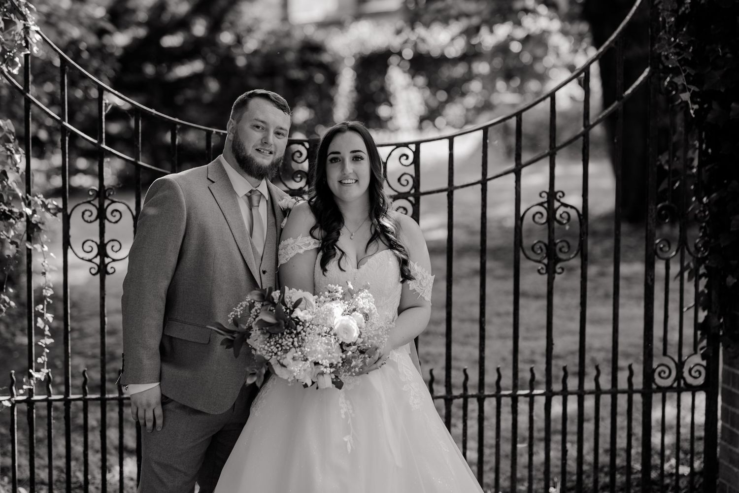 A romantic wedding couple shares an intimate moment by an ornate iron gate in a lush garden setting.