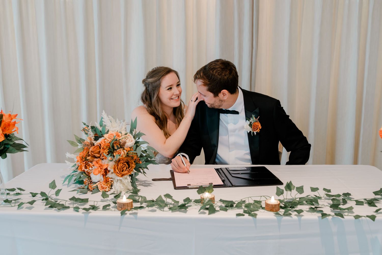 Wedding reception table decorated with orange floral arrangements and greenery against white curtain backdrop.