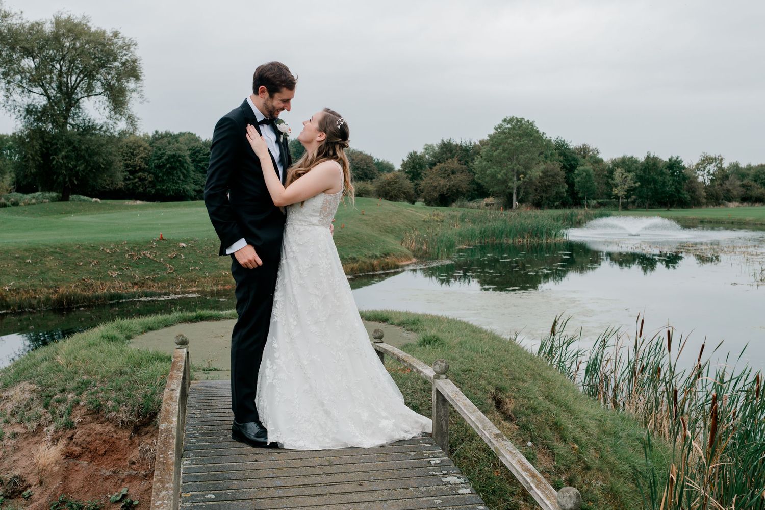 Bride and groom share a romantic kiss on a wooden bridge overlooking a serene pond at their outdoor wedding.