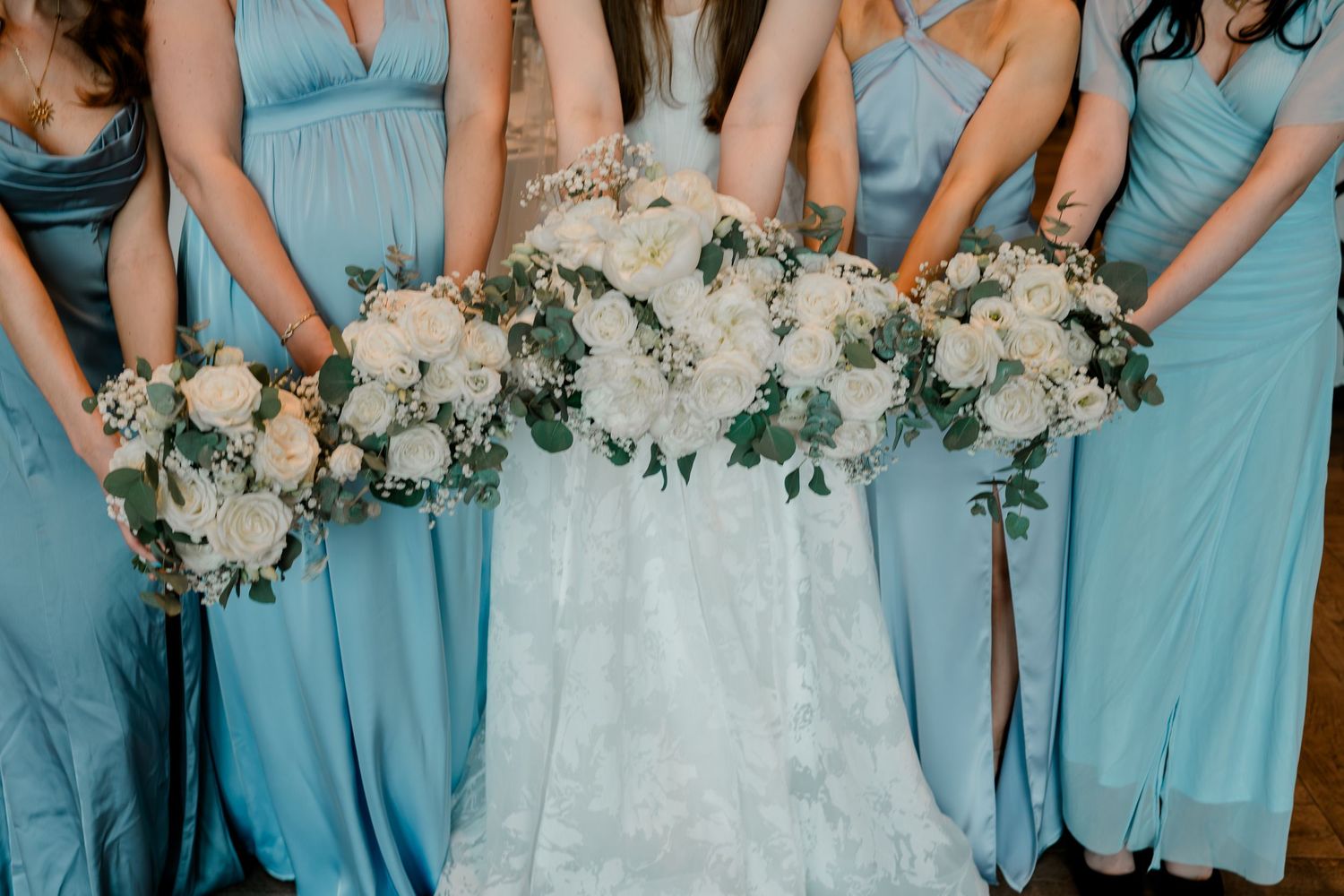 Bride and bridesmaids in light blue dresses holding white floral bouquets with greenery at a wedding ceremony.
