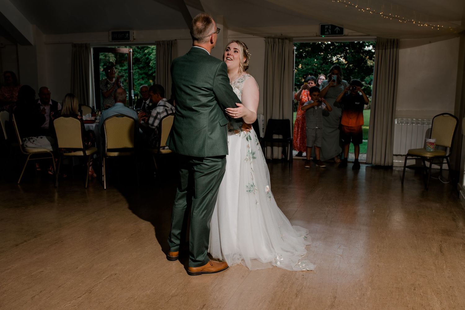Couple dancing in spacious reception hall with string lights overhead.