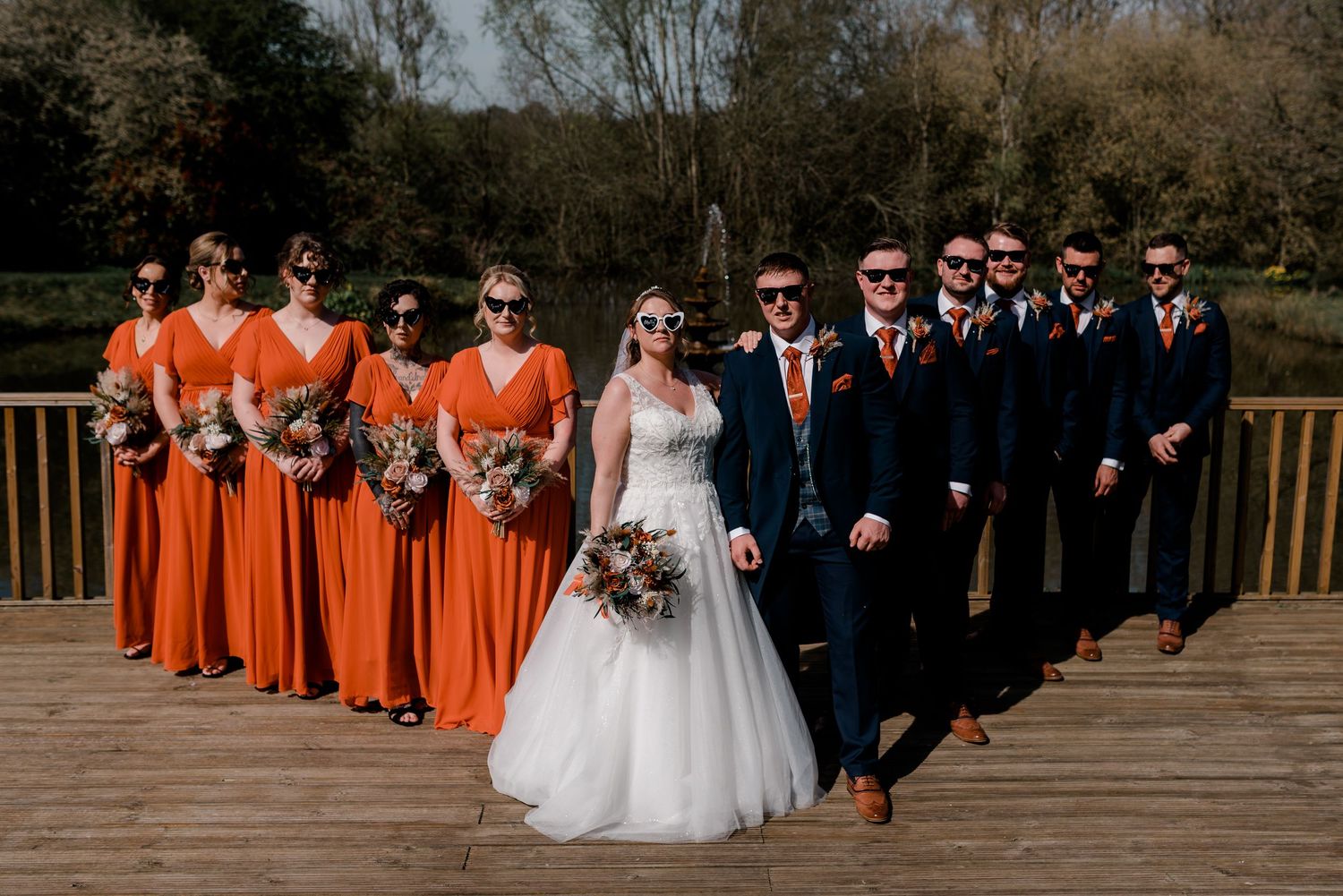 Wedding party poses on wooden deck with bridesmaids in bright orange dresses and groomsmen in dark suits.