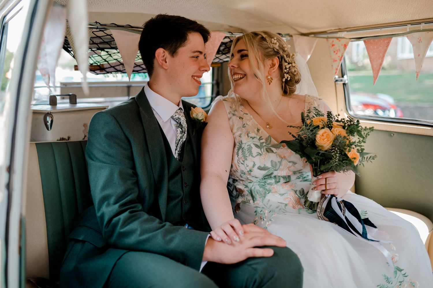 Smiling bride and groom sitting together in a vintage white wedding car with floral bouquet.