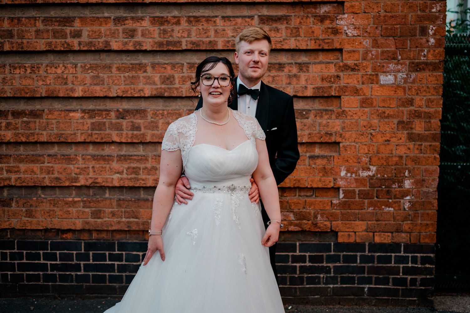 Bride and groom posing in front of brick wall, she in white A-line gown, he in black tuxedo.