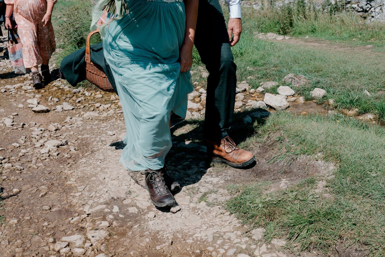 Close-up of wedding guests feet walking along a rocky dirt path, wearing boots and dress shoes outdoors.