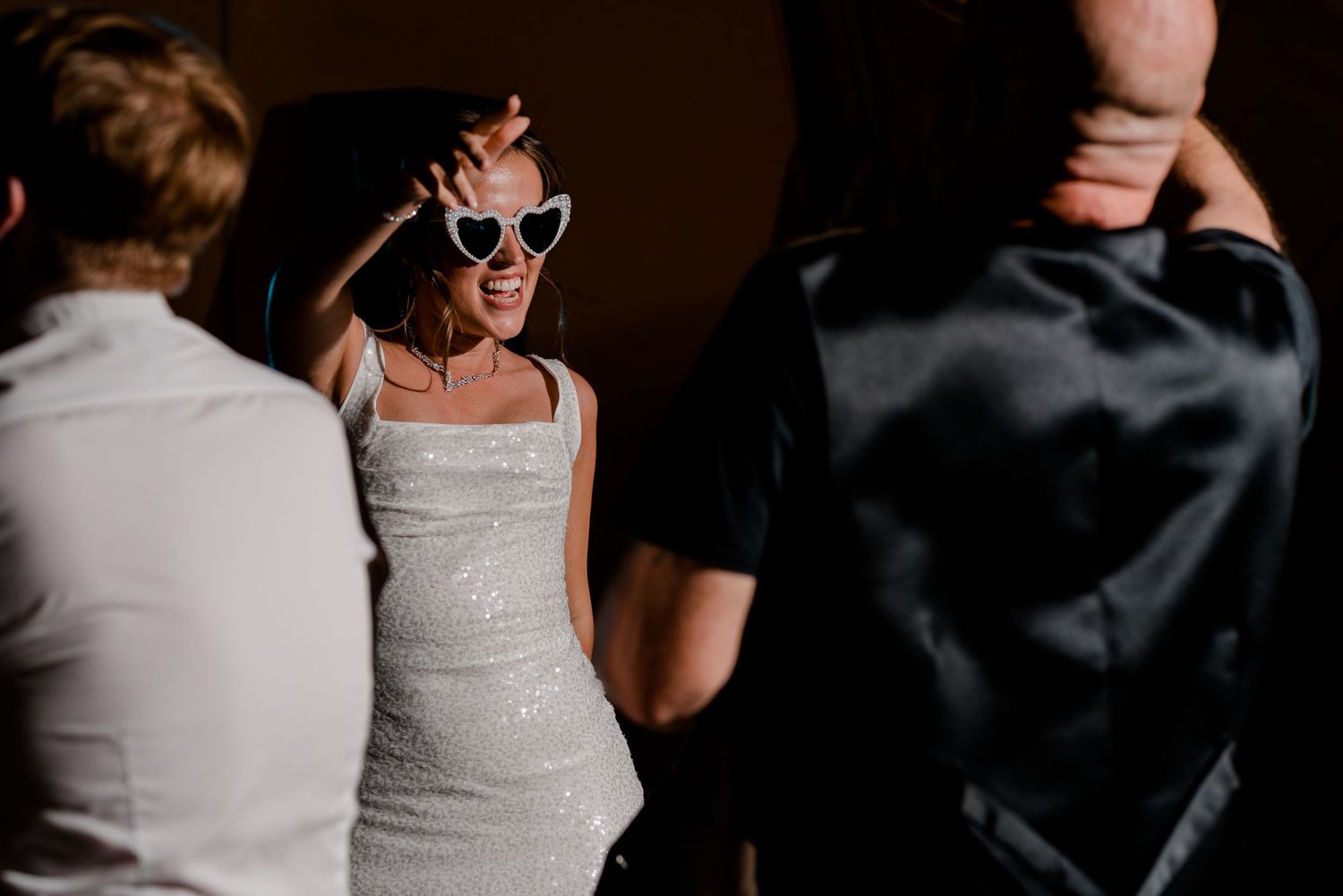 Woman in sequin dress wearing heart-shaped sunglasses dancing joyfully at a wedding reception.