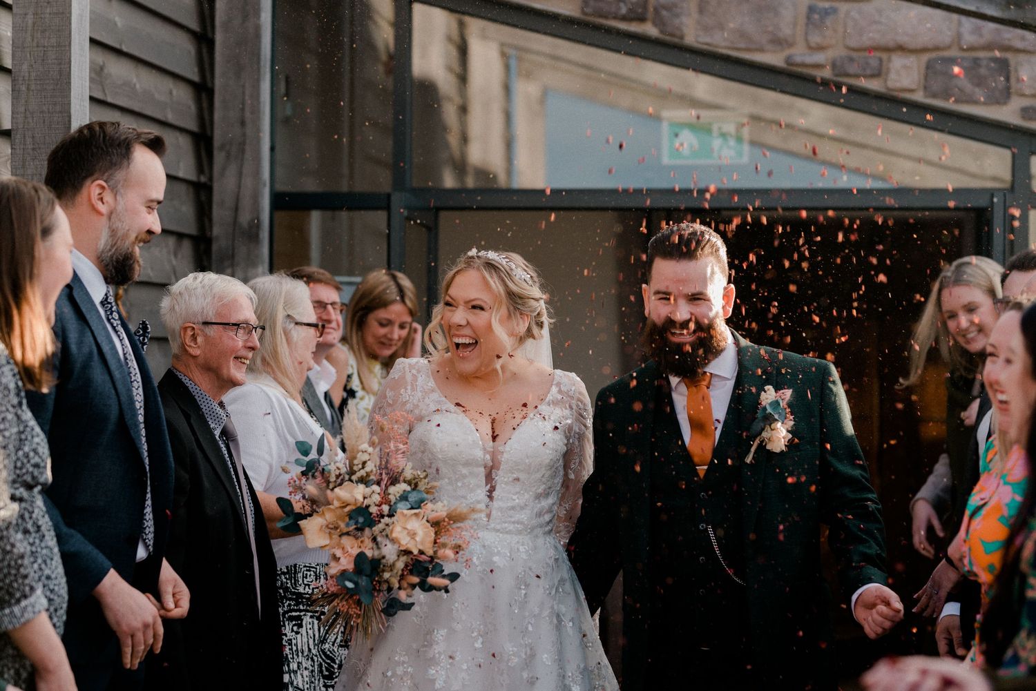 Happy bride and groom laughing during confetti exit outside rustic wedding venue surrounded by celebrating guests.