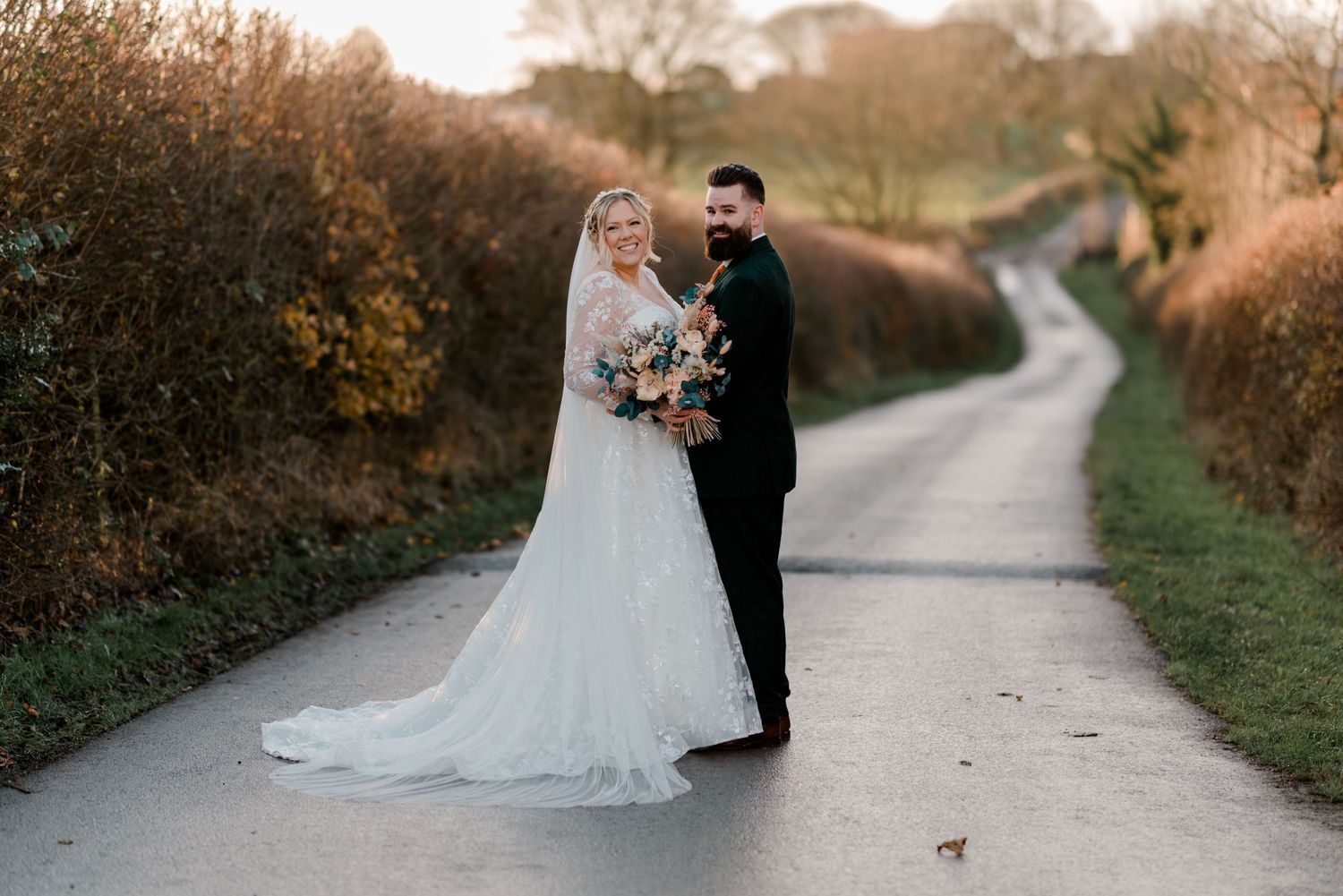 Couple in wedding attire standing on a country lane during autumn.