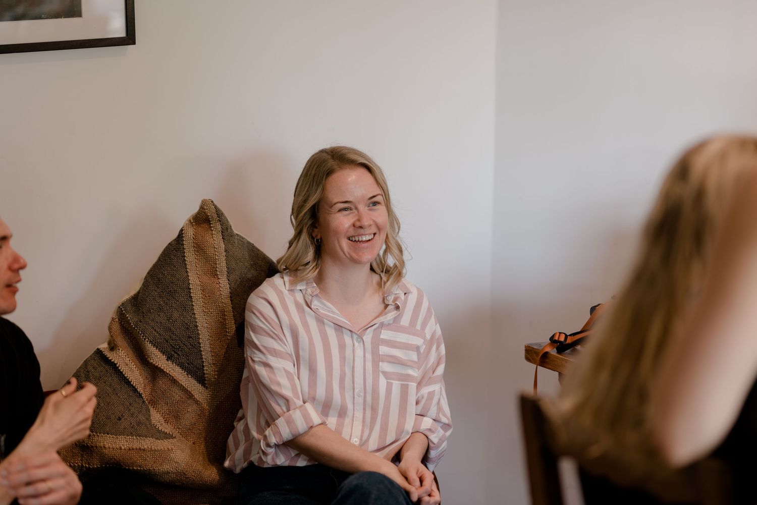 A person in a striped button-down shirt sits on a couch smiling during a casual indoor gathering.