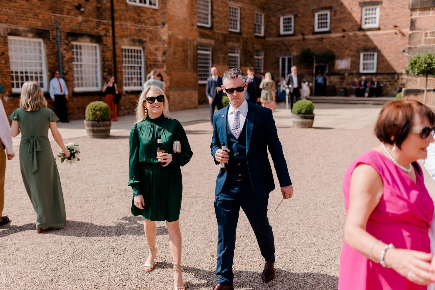 Wedding guests mingling in courtyard of historic red brick building with large windows.