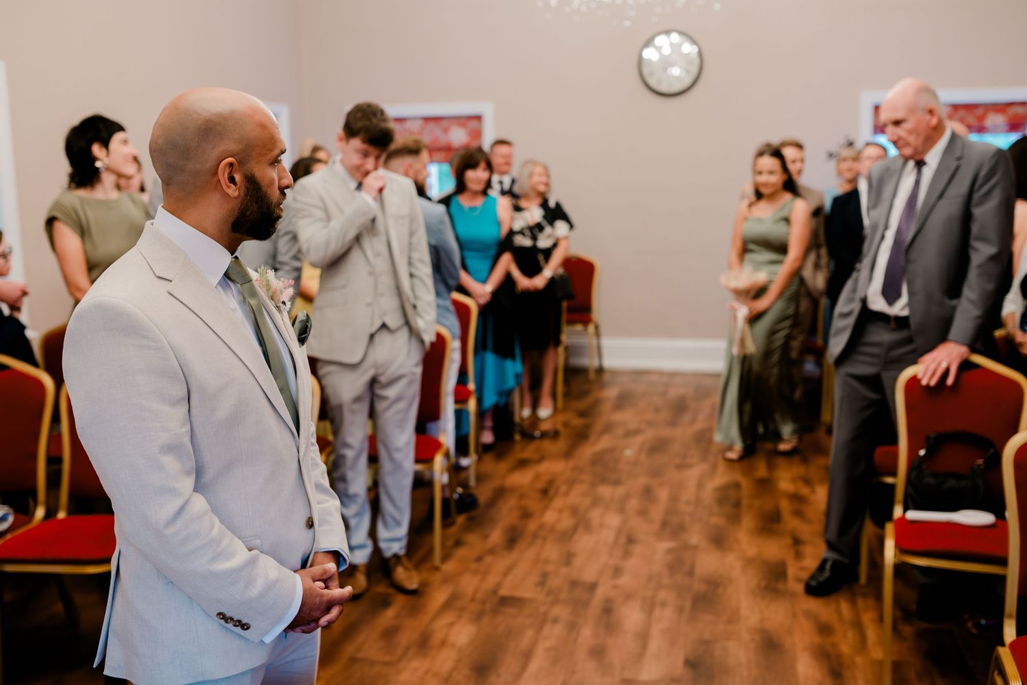 Wedding guests gather in an indoor reception venue with wooden floors and red chairs.
