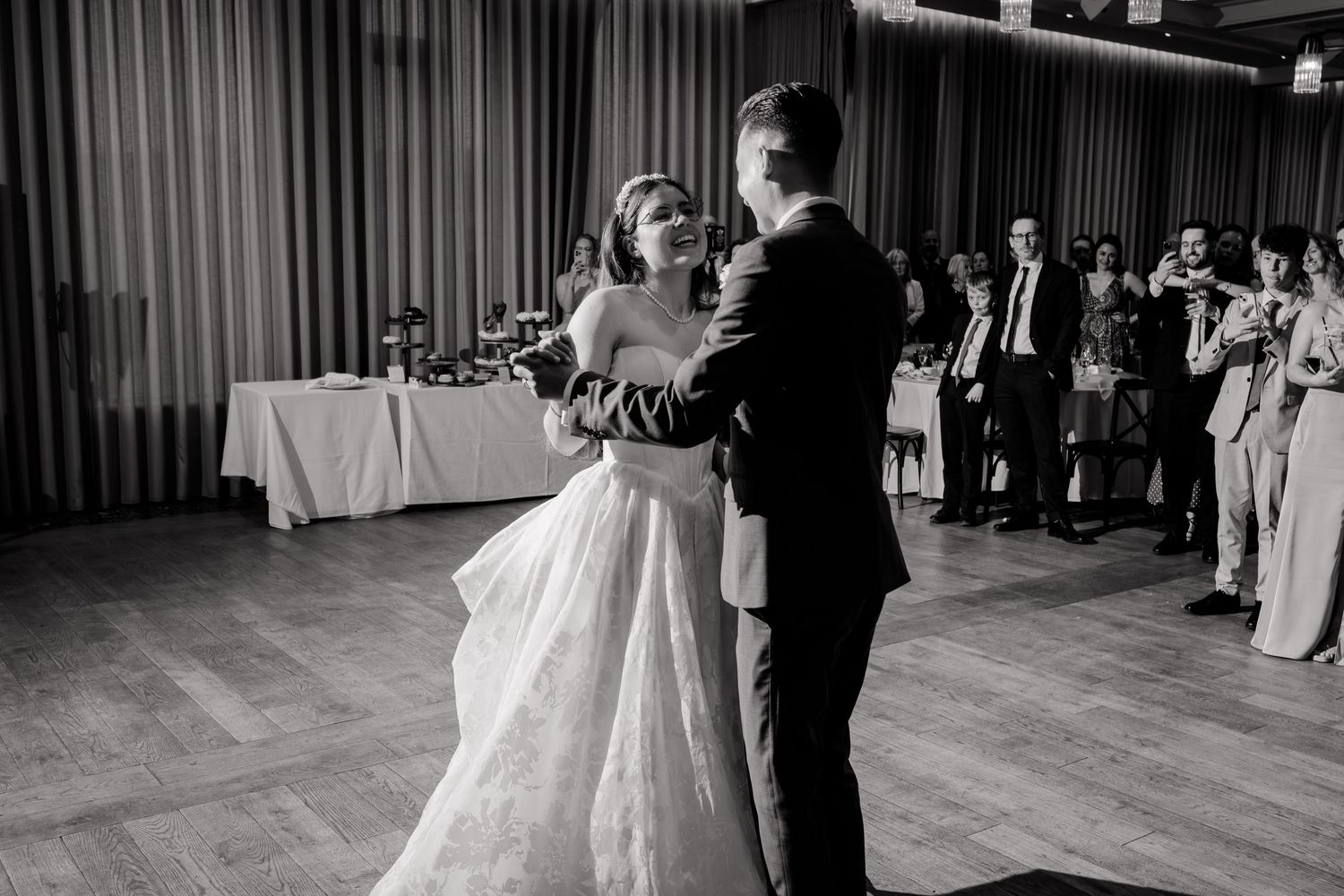 Bride and groom share their first dance in black and white photo with guests watching in background.